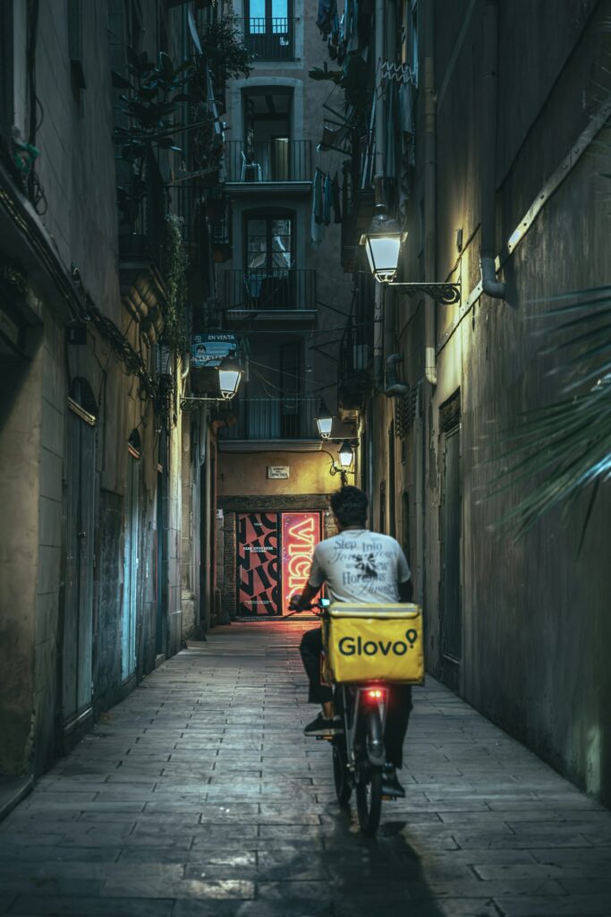 Delivery cyclist with a yellow box riding through a narrow stone alleyway